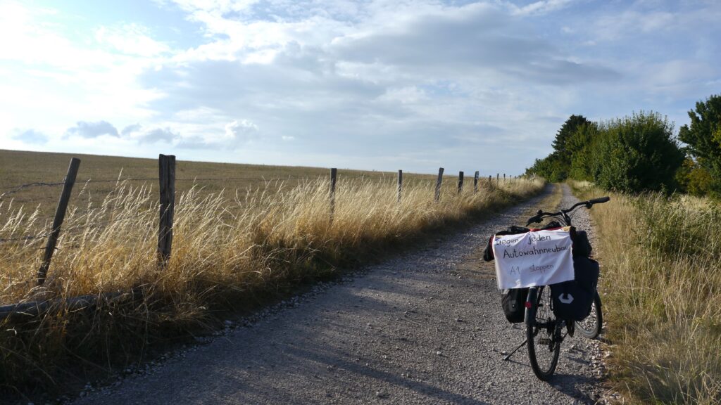 Auf dem Bild sieht mensche ein Fahrrad mit einem Transparent am beladenen Gepäckträger angebracht, auf dem steht "Gegen jeden Autowahnneubau - A1 stoppen". Das Fahrrad steht auf einem Feldweg. Links vom Weg ist ein vertrocknetes Feld.